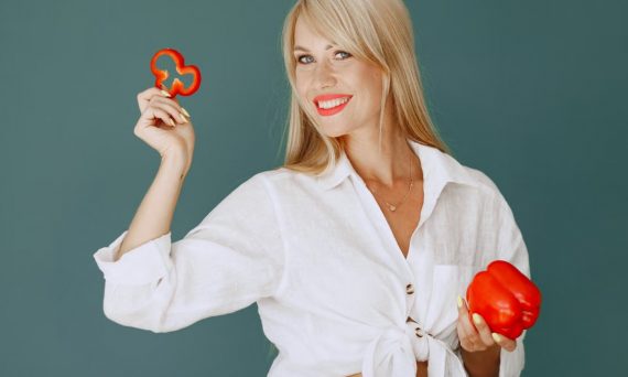 Blonde woman in white shirt smiling while holding a fresh red bell pepper slice.